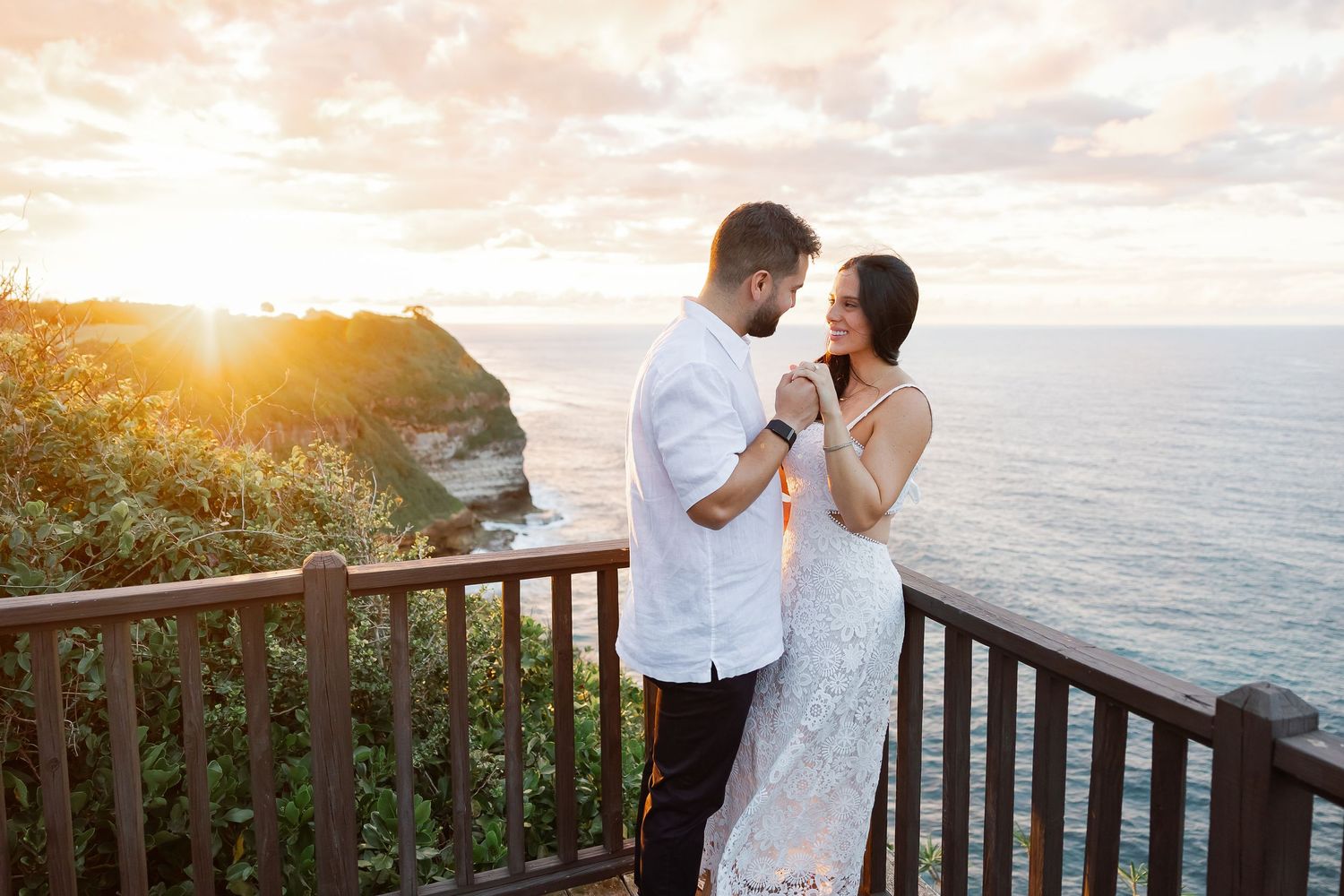 A couple embracing on a balcony overlooking the ocean during sunset.
