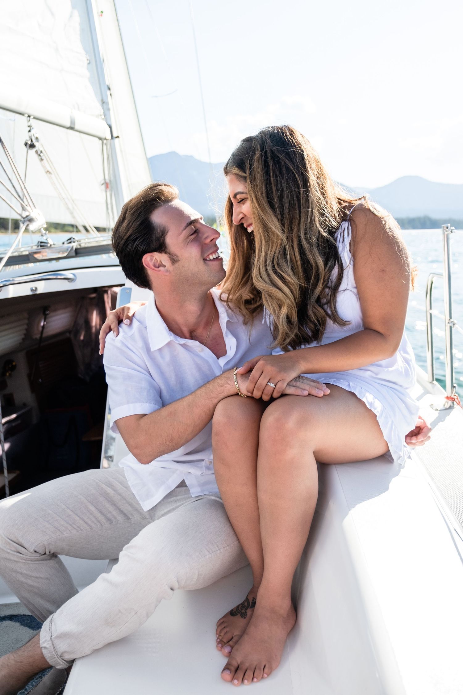 A happy couple shares a romantic moment on a white sailboat with mountains and blue water in the background.