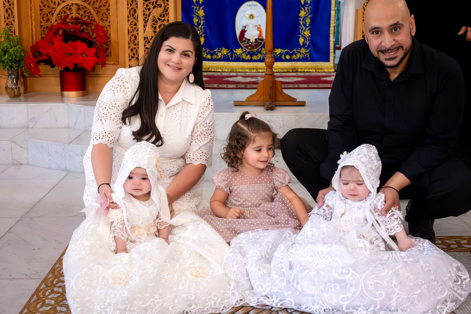 Family of four posing for a christening photo with two babies in white gowns and a toddler girl in a decorated church.
