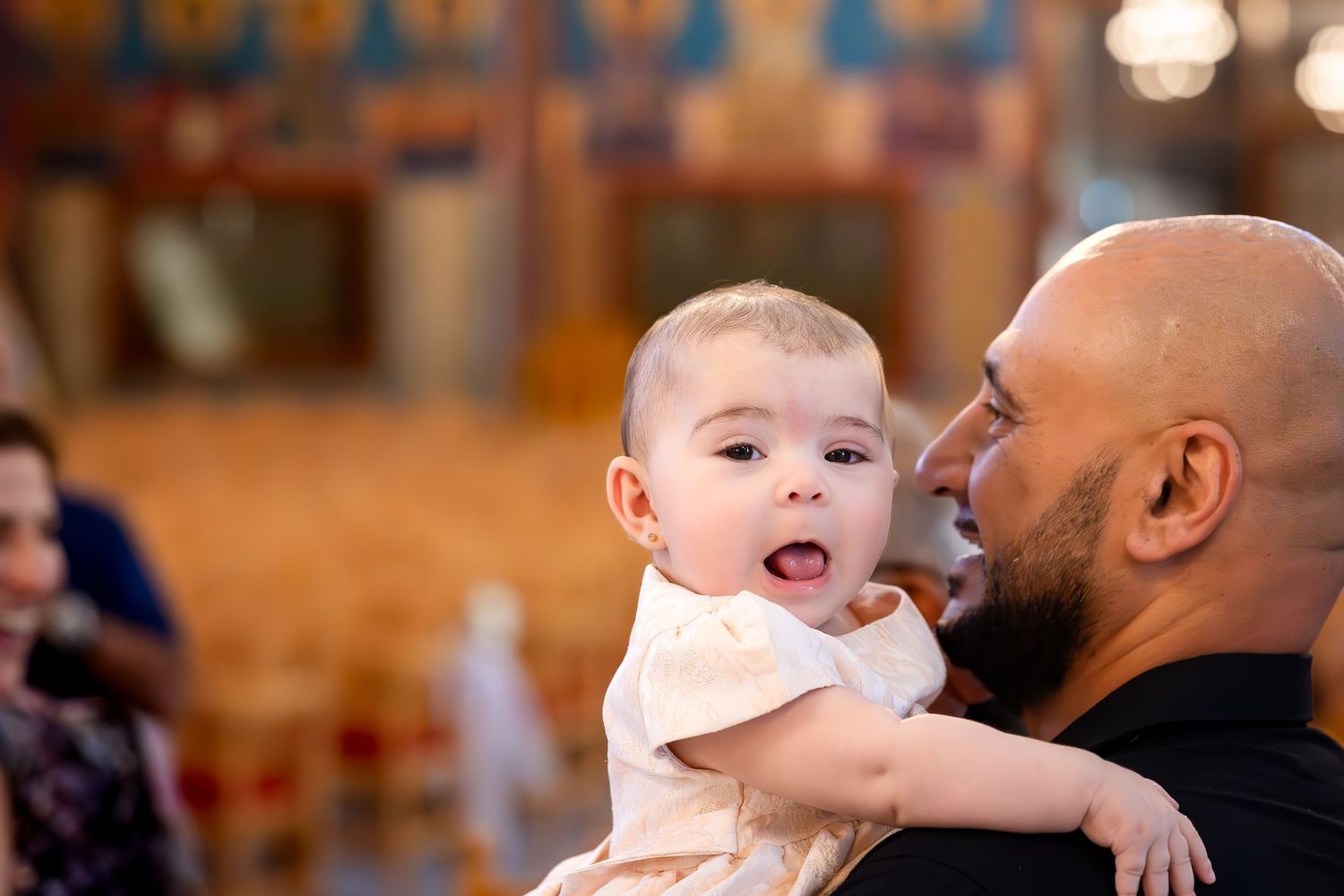 A bald man holds a cute baby in a white outfit during a church ceremony, with warm golden interiors in the background.