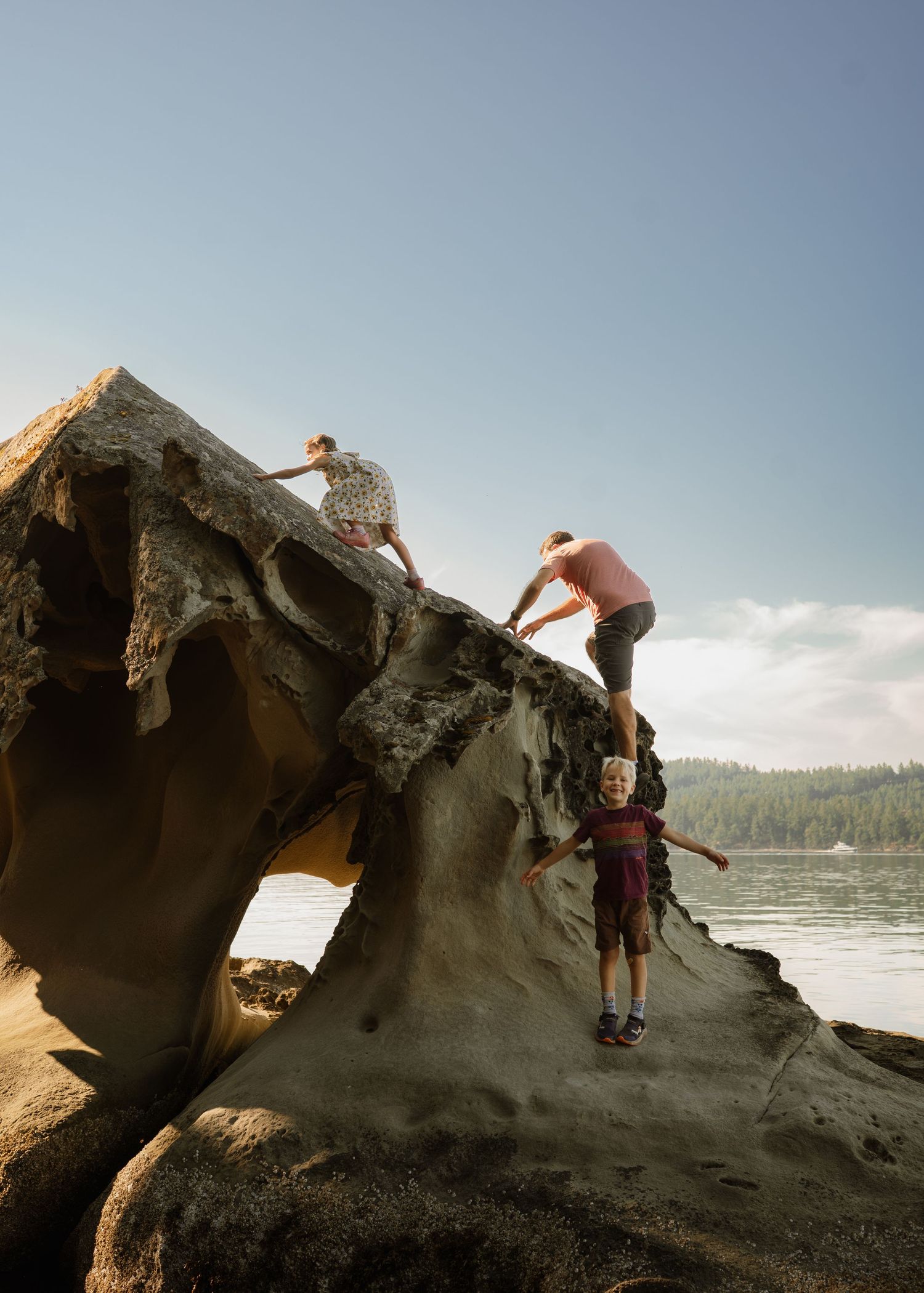 Family climbing a large sculpted rock formation on a scenic lakeside shore with forested hills in the background.