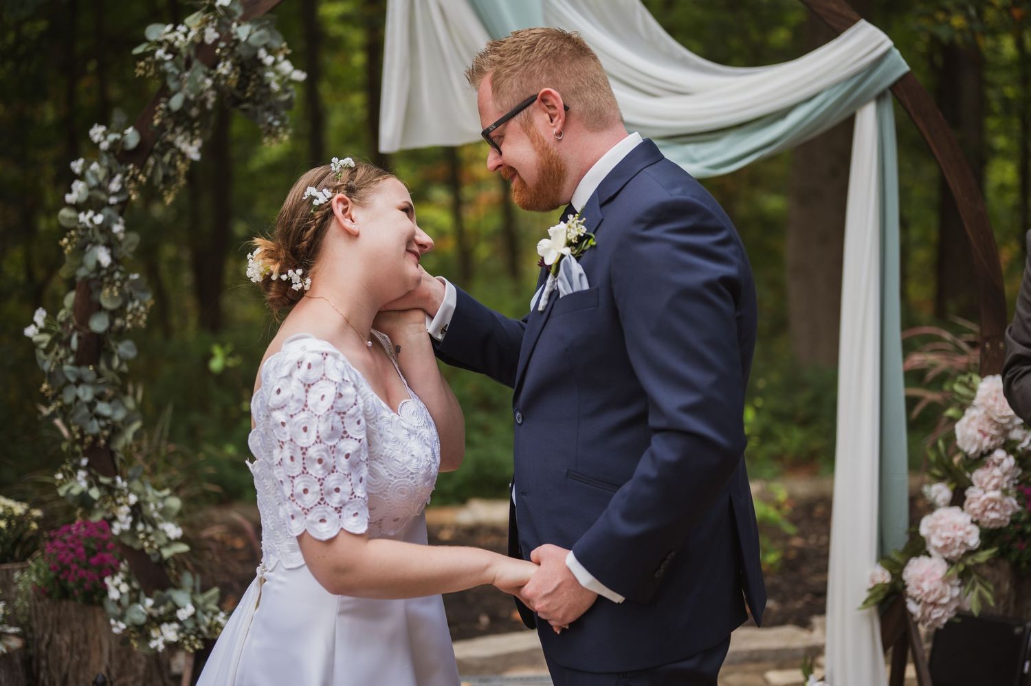 A joyful couple shares a candid moment during their outdoor wedding ceremony under a floral arch and white draping.
