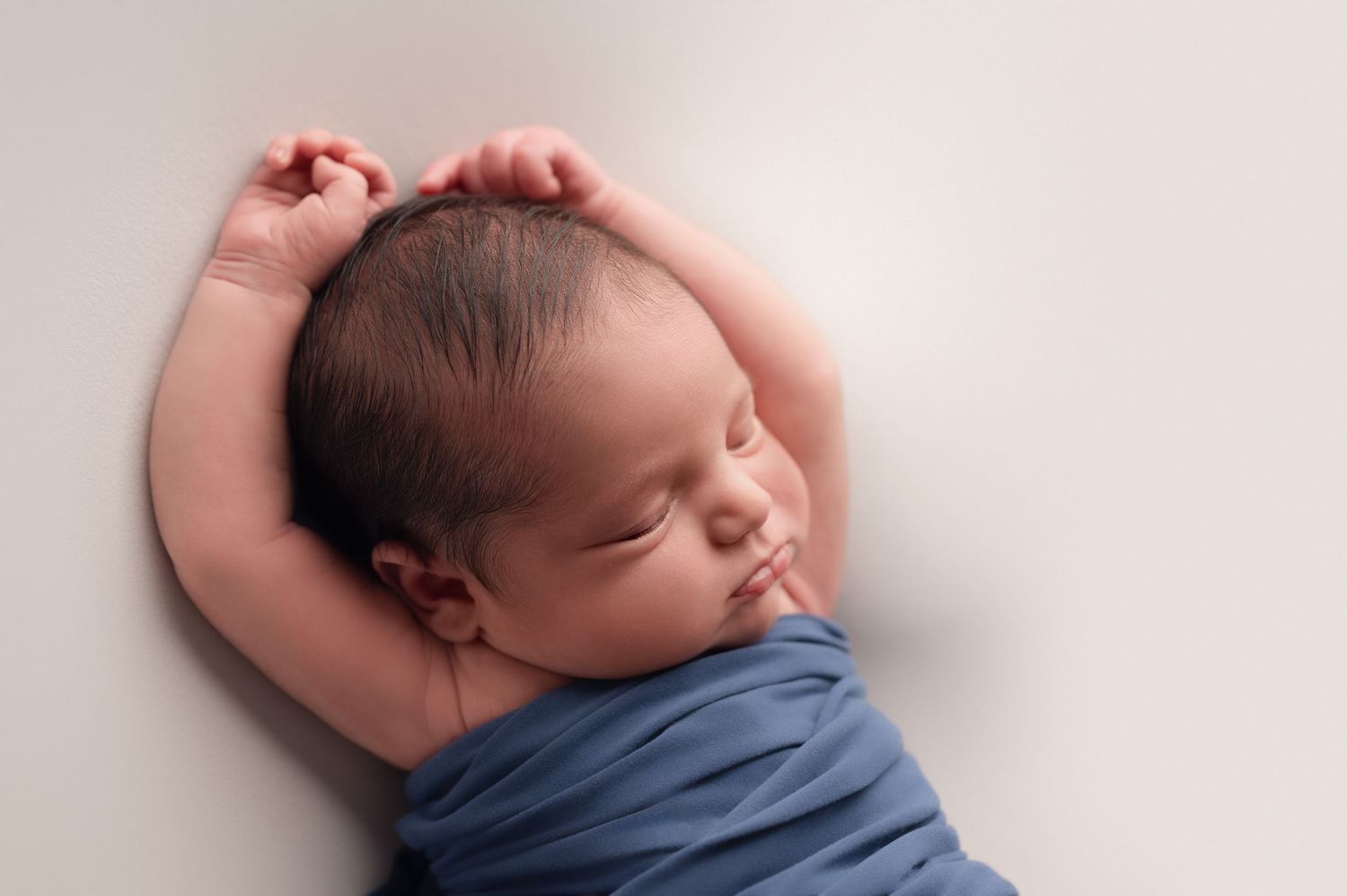 A sleeping newborn baby wearing a blue wrap stretches their arms above their head while resting on a soft blanket.