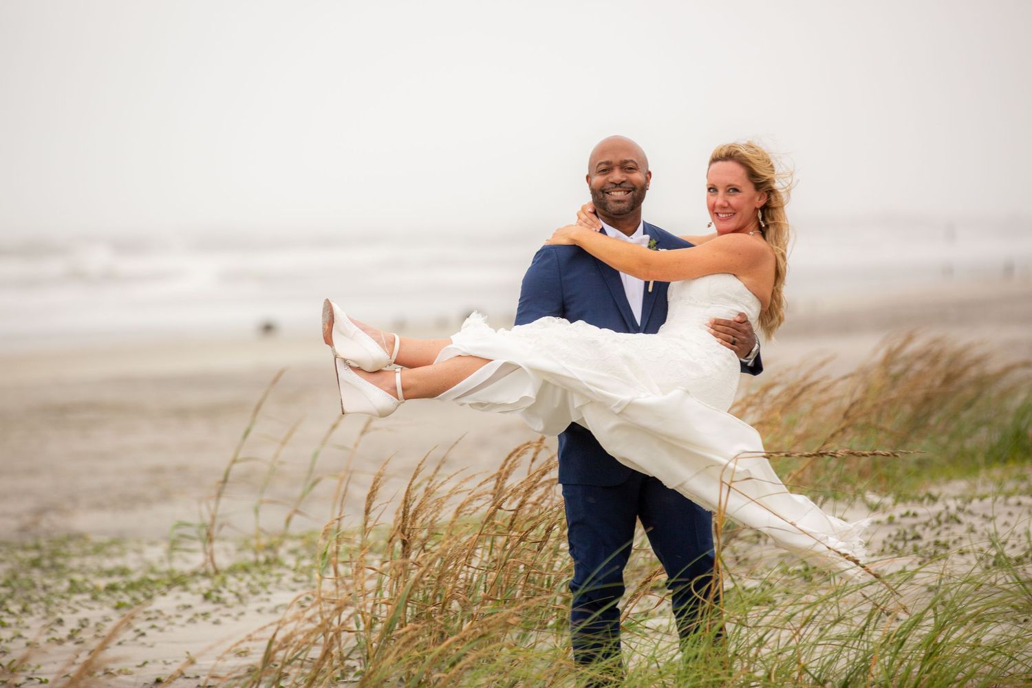 A couple shares a romantic moment on a misty beach as tall grass sways in the foreground.