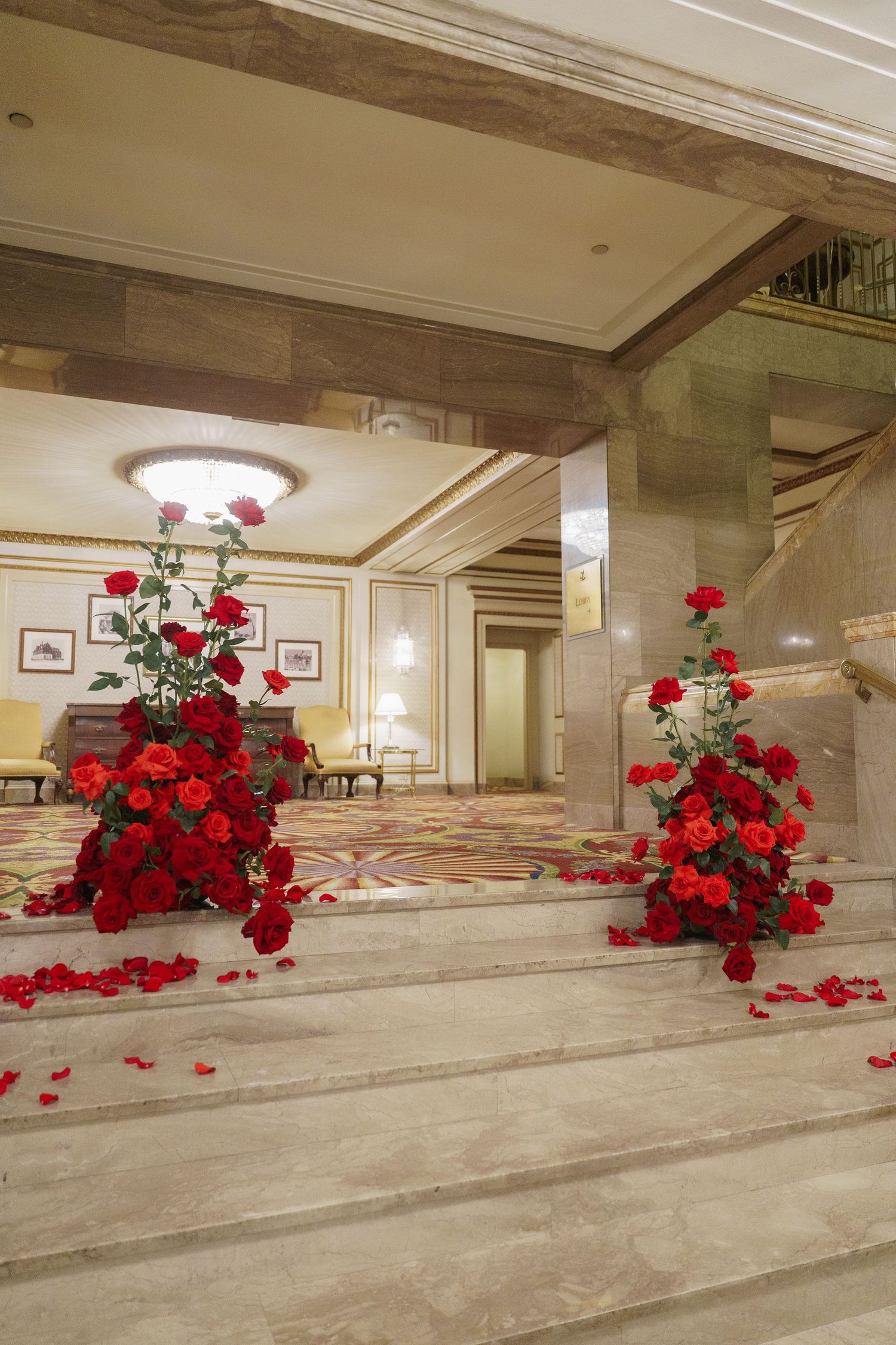 Red rose arrangements cascade down elegant marble steps in a luxurious hotel lobby with cream-colored walls and lighting.