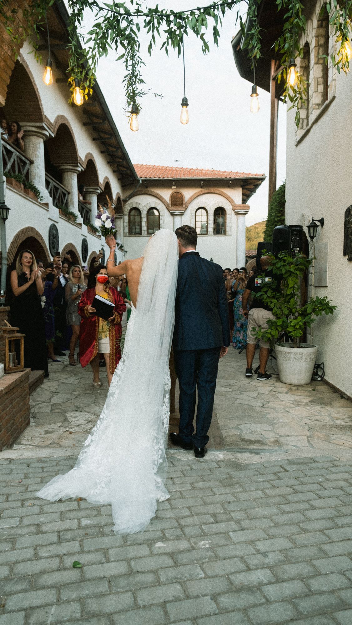 Wedding couple walks down an elegant stone courtyard with white columns and hanging greenery at sunset.