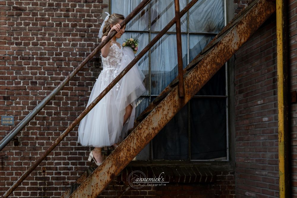 A bride in a white wedding dress poses against a rustic brick wall and metal staircase in an urban setting.