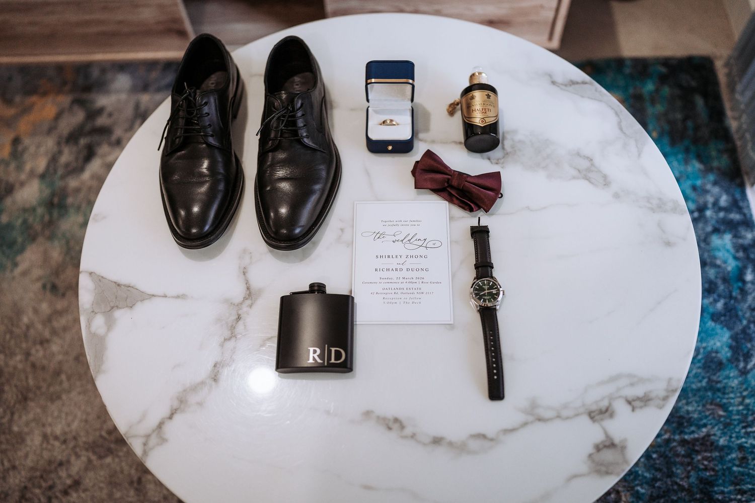 Wedding accessories including shoes, cologne, watch, and rings arranged on white marble surface.