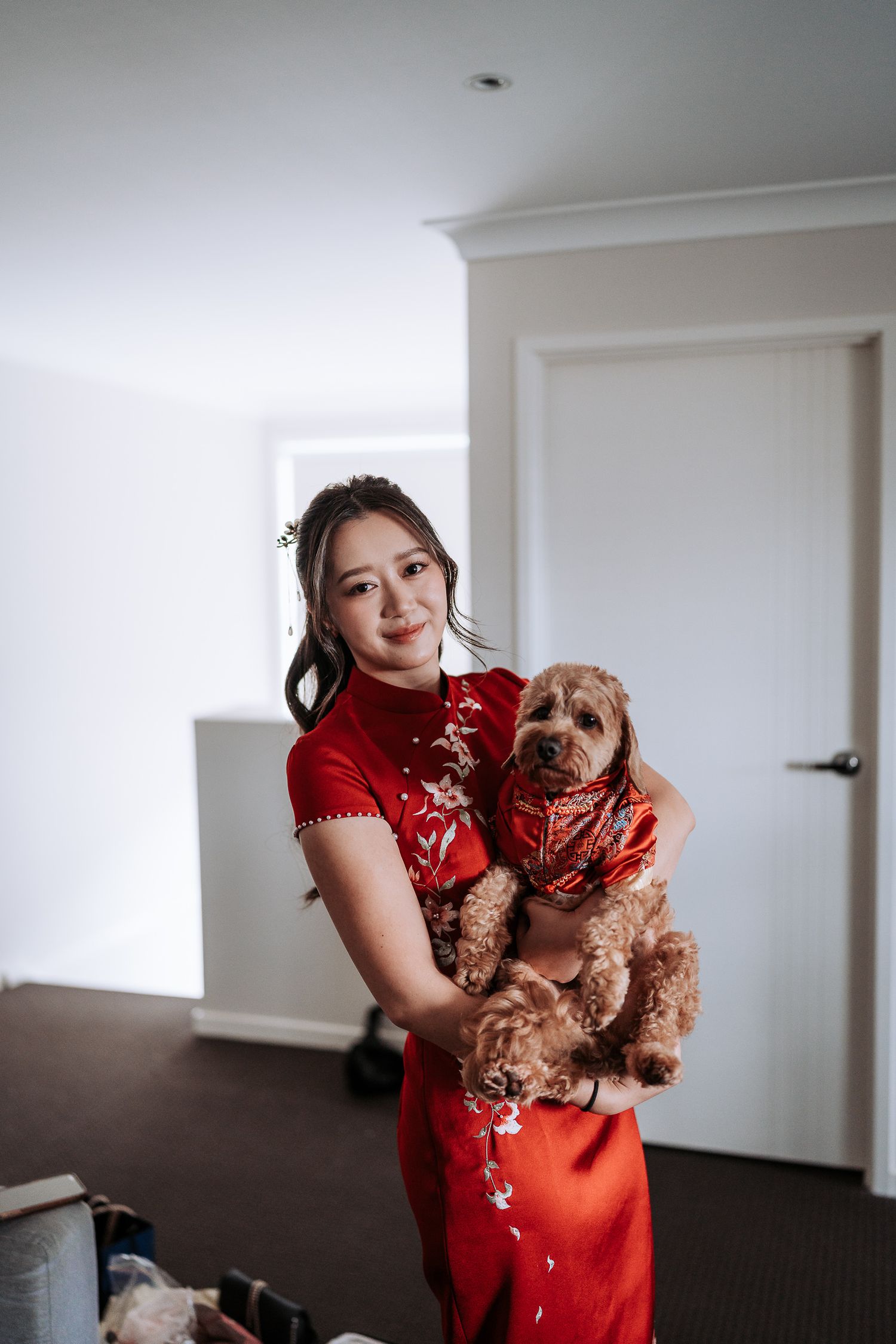 A person in a red traditional Chinese qipao dress holds a small brown dog wearing matching attire in an indoor setting.