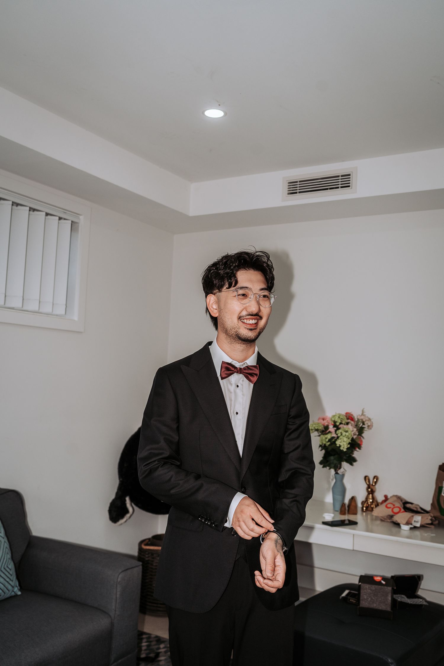 A person in a black suit and red bowtie stands in a room with white walls and a vase of flowers on a table.