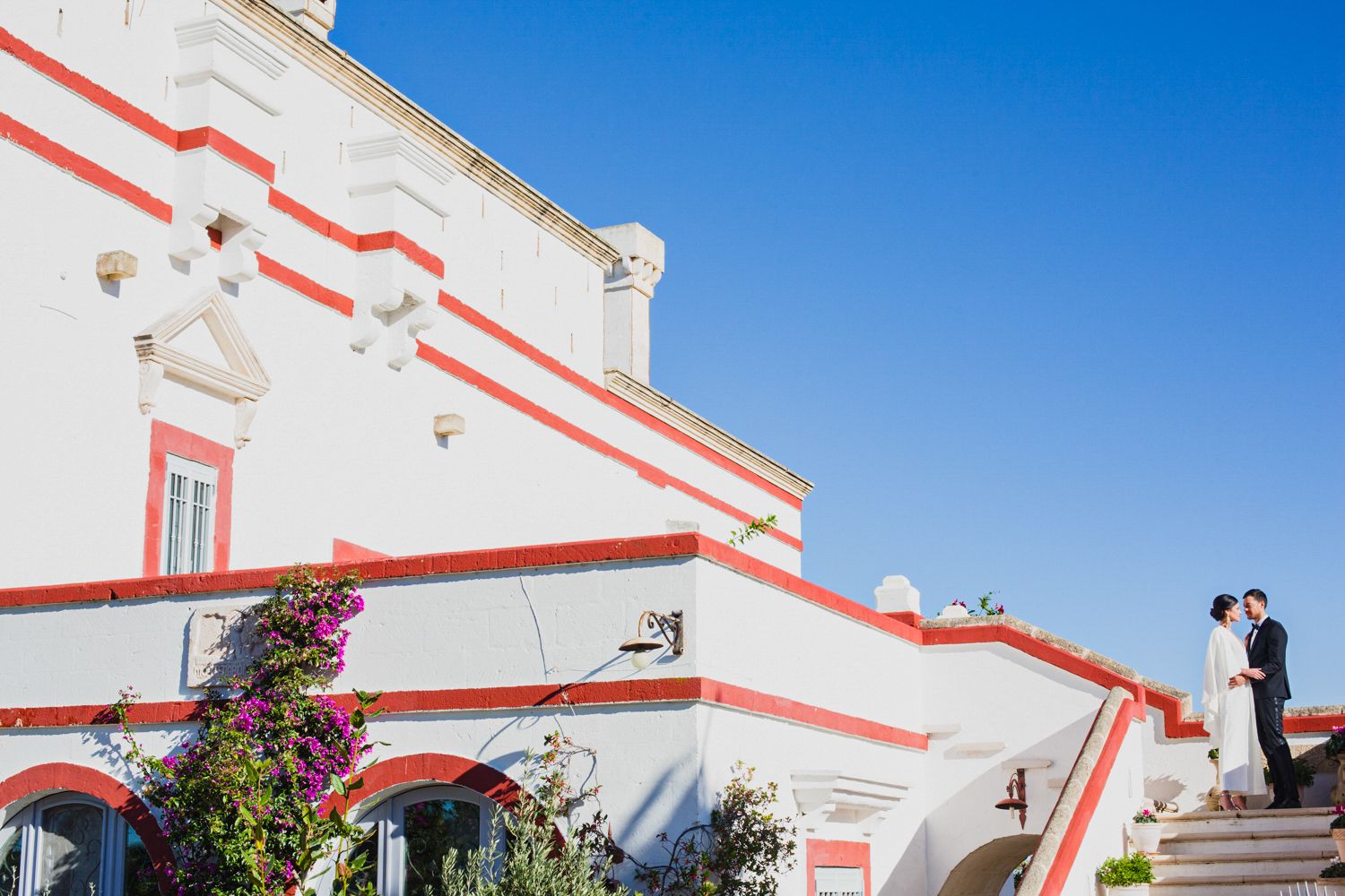 Bride and groom standing on white building with red trim under blue sky at outdoor wedding venue.