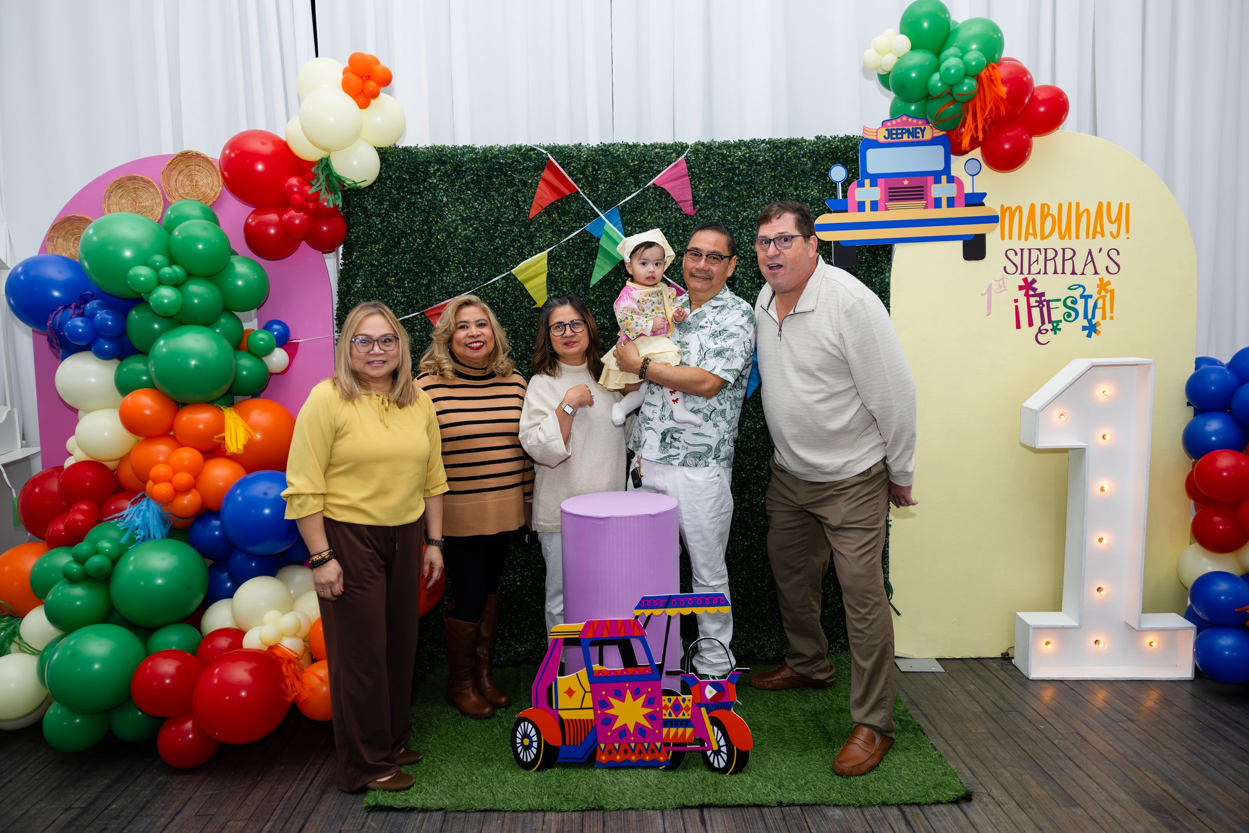 Family celebrating a first birthday party with colorful balloon decorations and a festive backdrop.