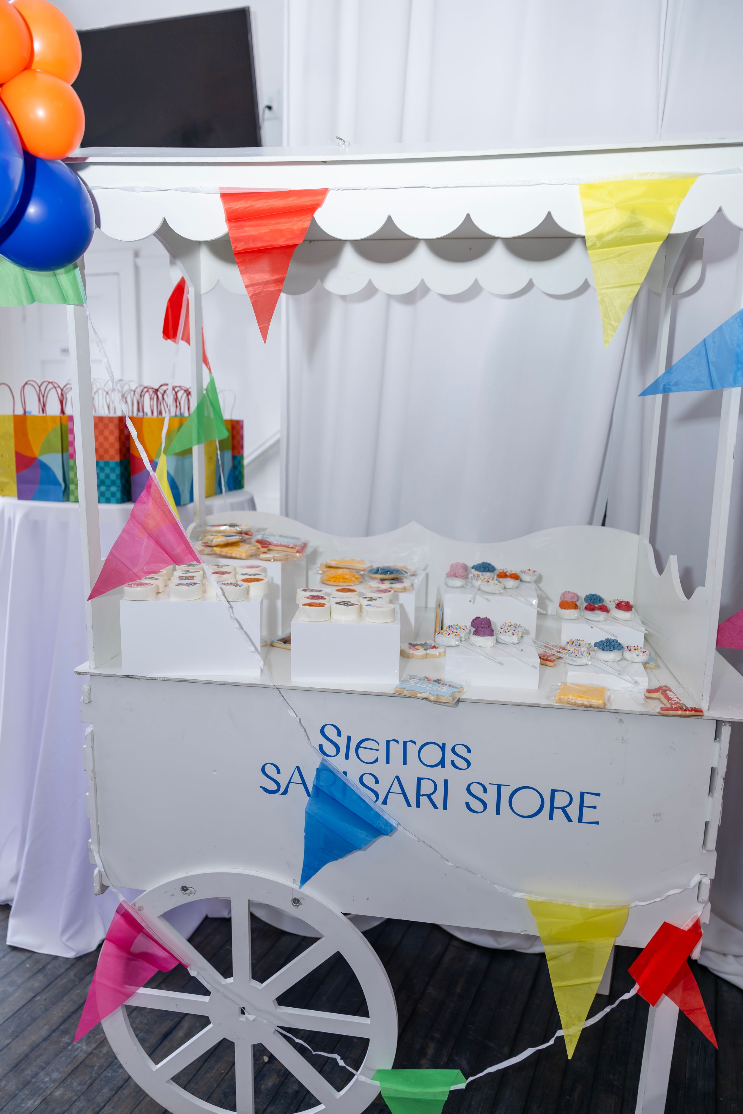 White cart vendor display labeled Sierras Sari-Sari Store with colorful bunting and treats at a fiesta party.