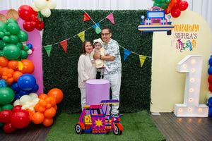 Family posing at a colorful first birthday party with balloon arch, green hedge wall, and festive decorations.