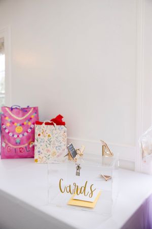 Clear acrylic cards box with gold lettering on a white table with colorful gift bags at a birthday party.