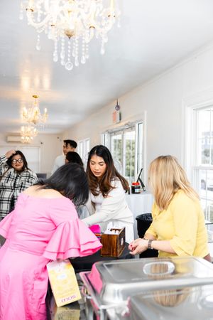 Women gather around a counter in an elegant white cafe adorned with crystal chandeliers.