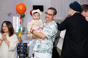 Man in Hawaiian shirt holds baby in graduation cap at festive indoor party with balloons and guests.