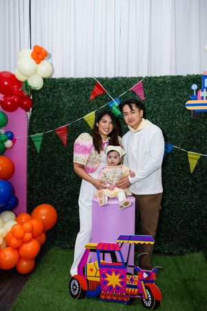 Family of three poses at colorful Filipino-themed first birthday party with jeepney prop and balloons.