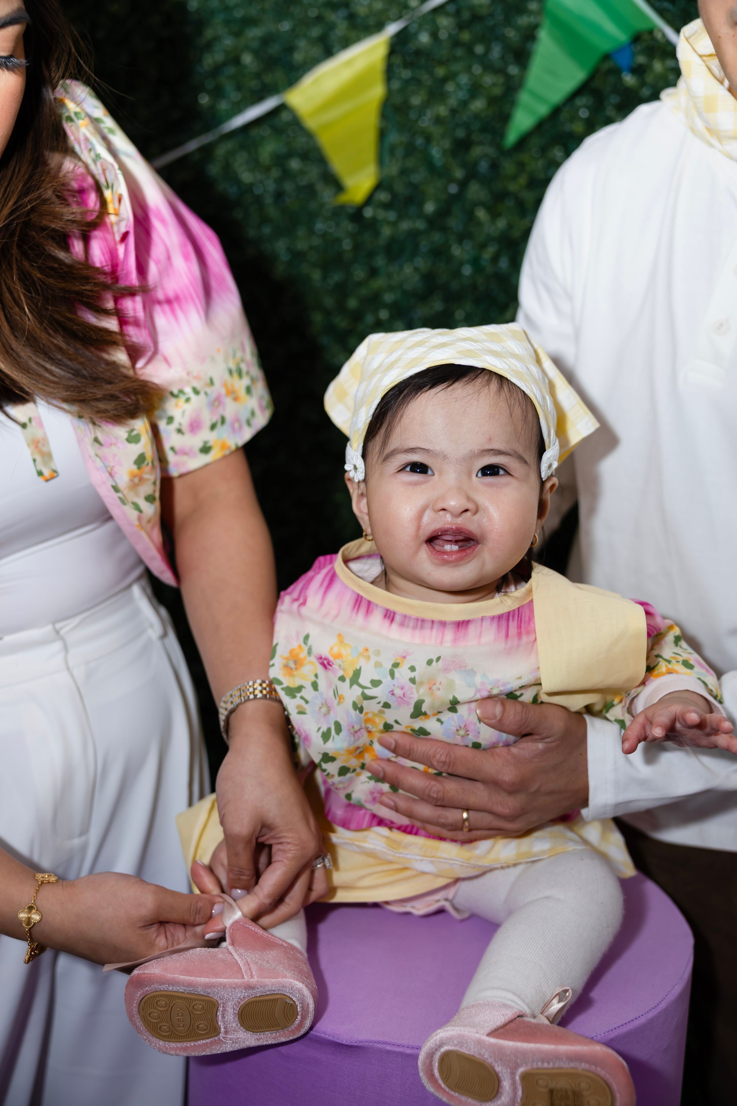 Baby girl in floral dress and yellow bonnet smiles at first birthday party with colorful decorations.