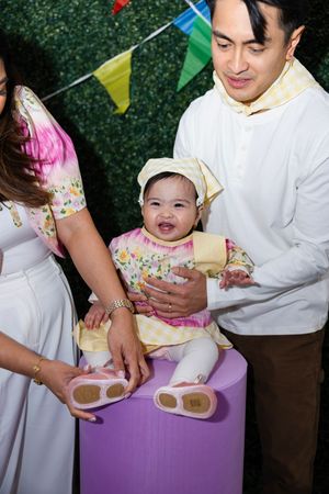 Happy baby girl in floral dress and yellow hat sits on purple pedestal with smiling parents at birthday party.