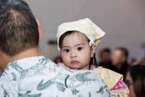 Adorable baby wearing yellow checkered hat rests on adult's shoulder at indoor celebration event.