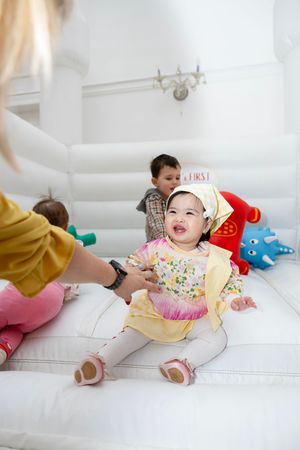 Smiling baby girl in floral dress and white hat sitting in a white bounce house at her first birthday party.
