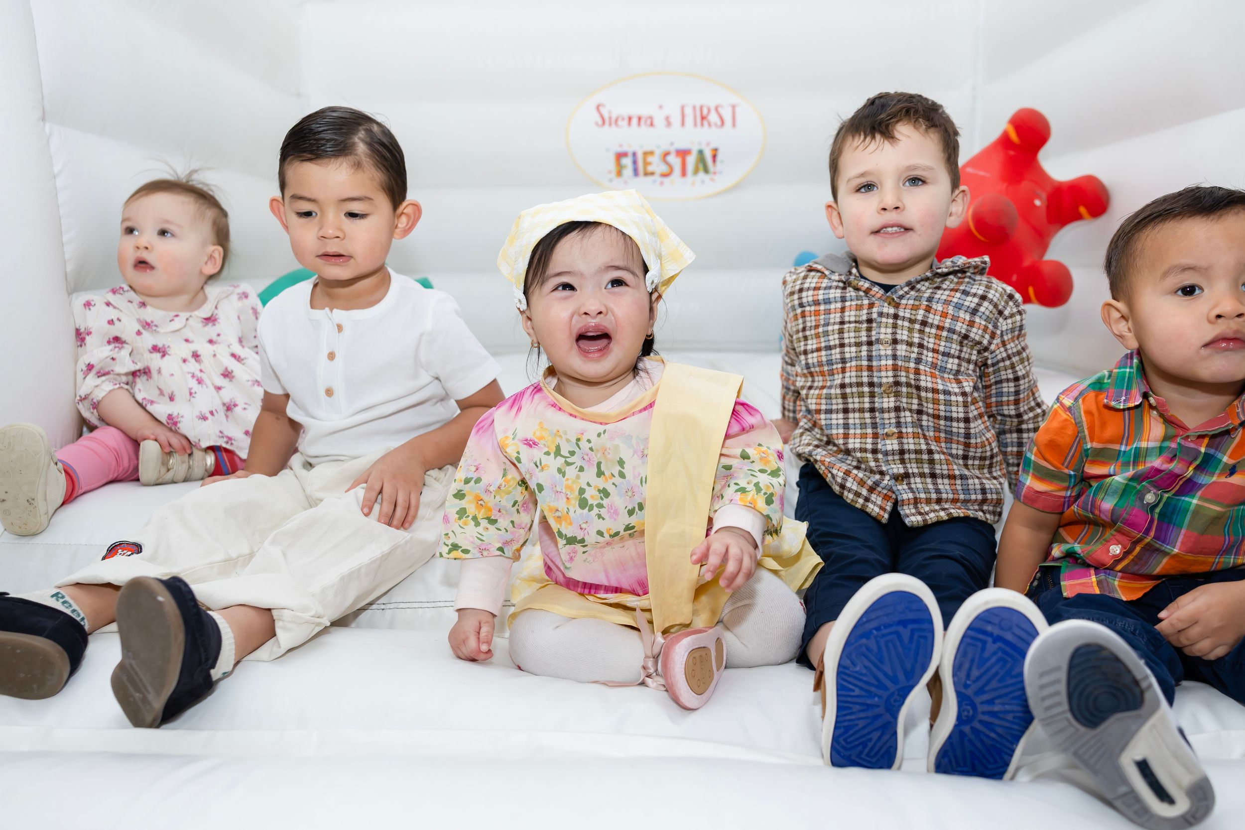 Five babies and toddlers sitting together at a first fiesta birthday party celebration in a white bounce house.