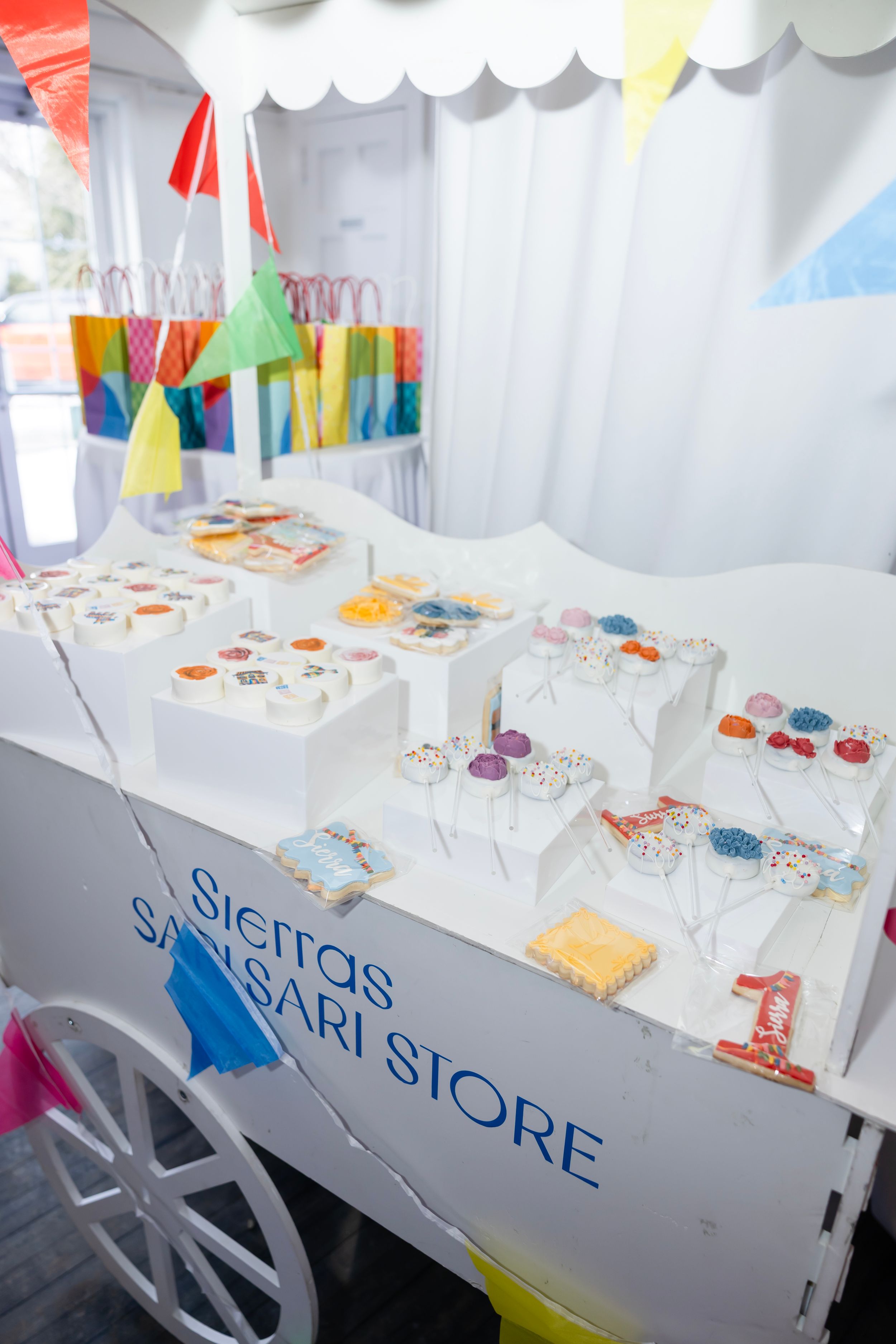 White cart display for Sierras Sansari Store featuring colorful treats, cake pops, and decorated boxes at a festive party.