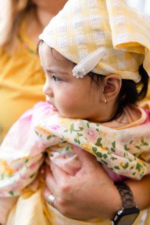 Close-up of infant in floral outfit and yellow hat with white flower clip being held at party.