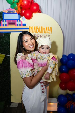 Smiling mother holding her baby girl in matching floral outfits at a Filipino-themed first birthday party.