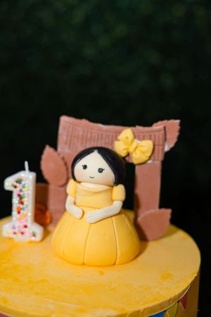 Close-up of a yellow first birthday cake topper featuring a handmade fondant girl doll with a number one candle.