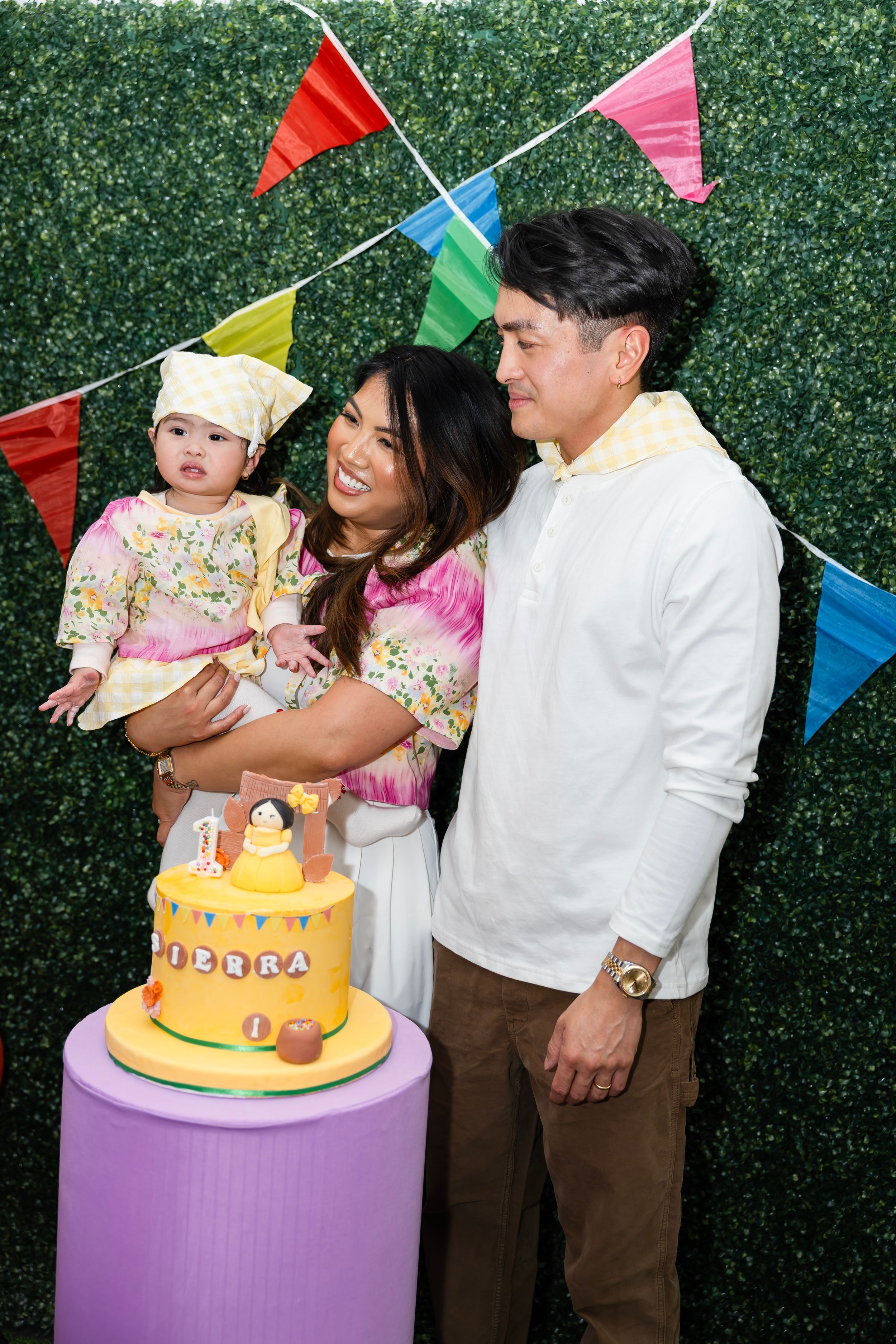 Family portrait of parents and baby girl with a yellow Sierra first birthday cake at a fiesta-themed party.