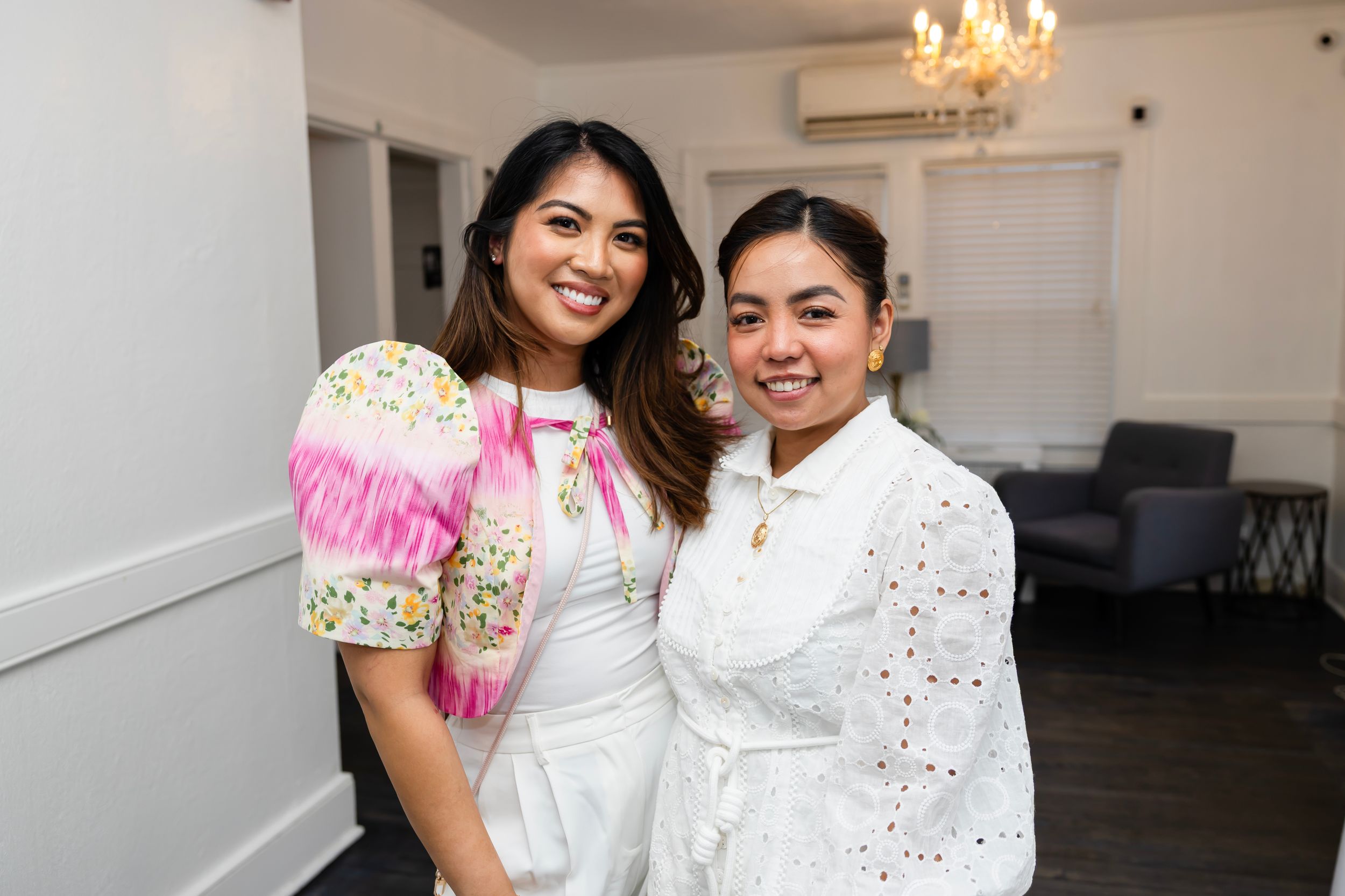 Two women smiling together at an indoor event, wearing elegant white and floral outfits in a bright, stylish venue.