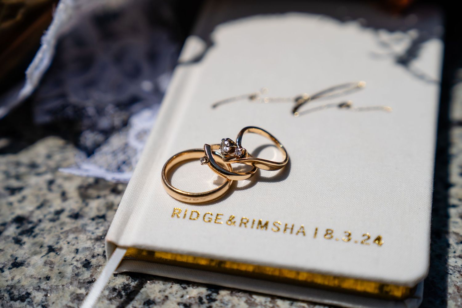 Gold wedding rings rest on a white personalized wedding journal engraved with Ridge & Rimsha 18.3.24.