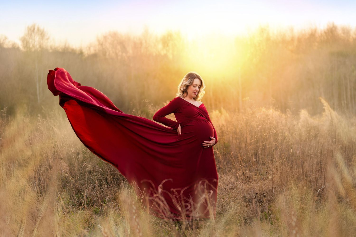 Pregnant woman in flowing red gown poses in golden field at sunset, dress billowing in wind.