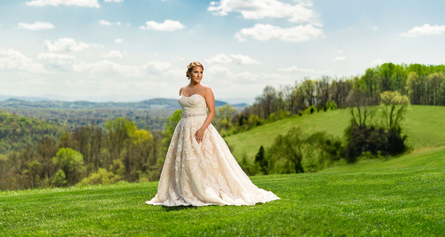 Bride in elegant white ball gown standing on lush green hillside with scenic mountain landscape backdrop on sunny day.