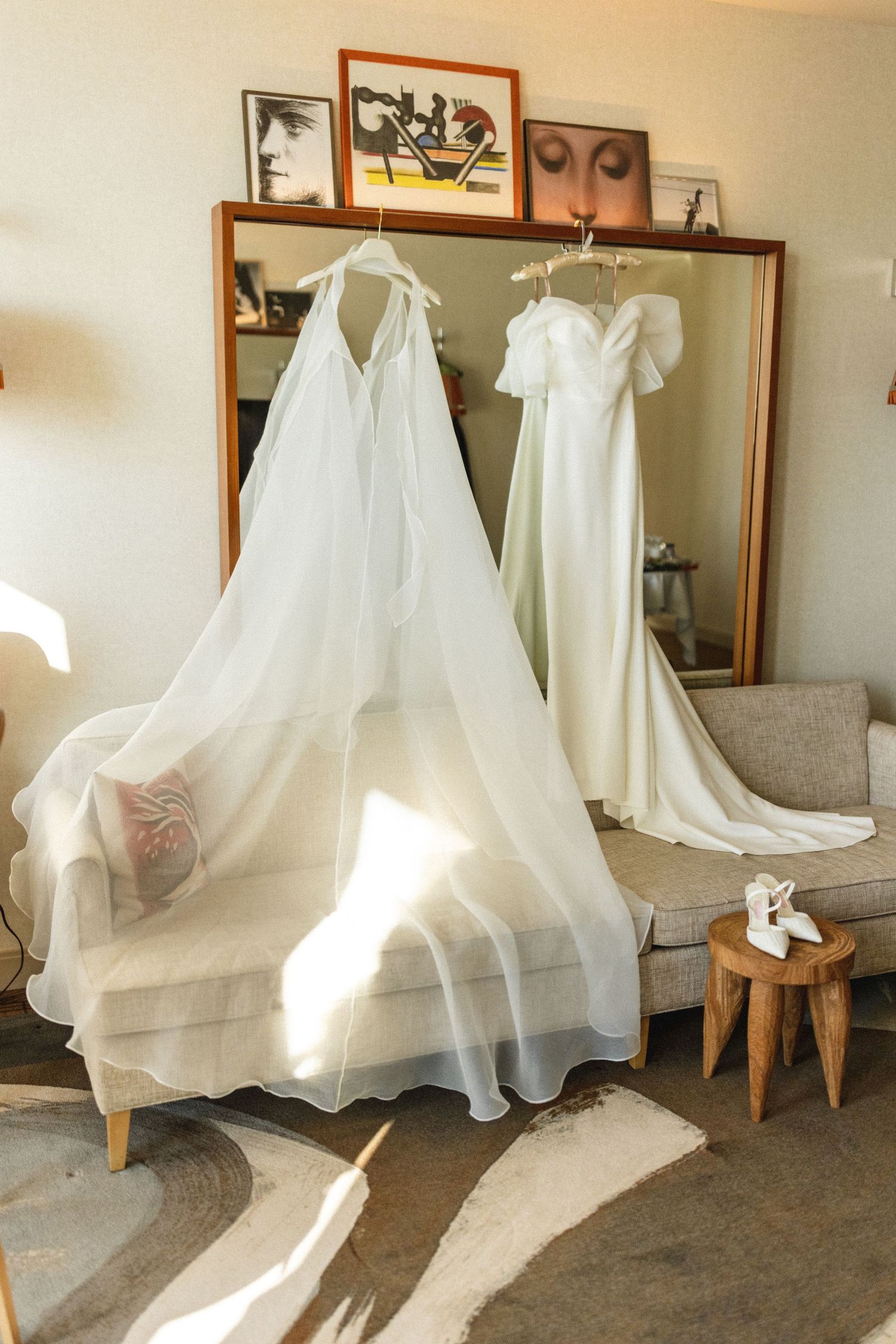 Two white wedding dresses hanging in a sunlit hotel room with a large mirror and vintage artwork.