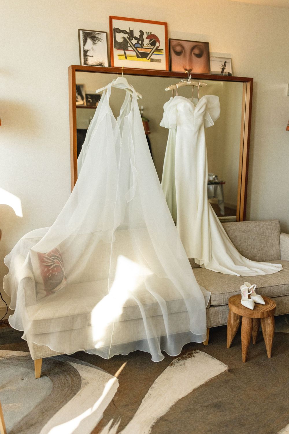 Two white wedding dresses hanging in a sunlit hotel room with a large mirror and vintage artwork.