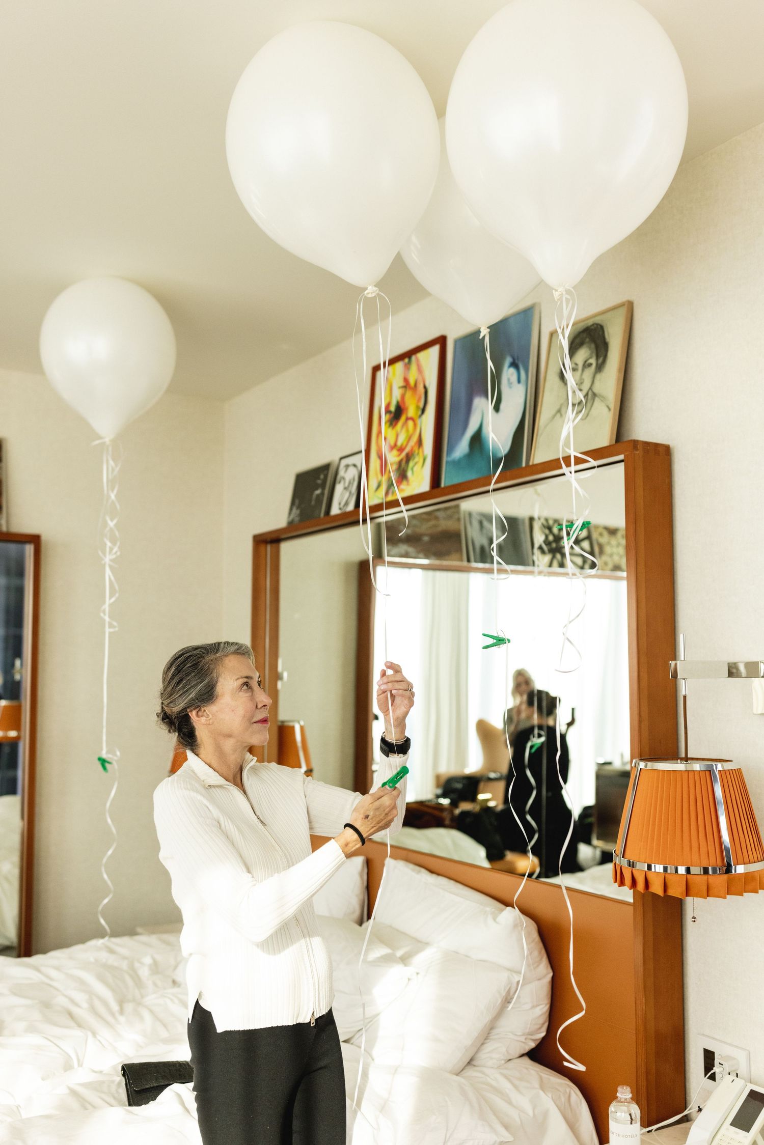 An older woman arranges large white balloons in a decorated bedroom with a mirror and framed photos.