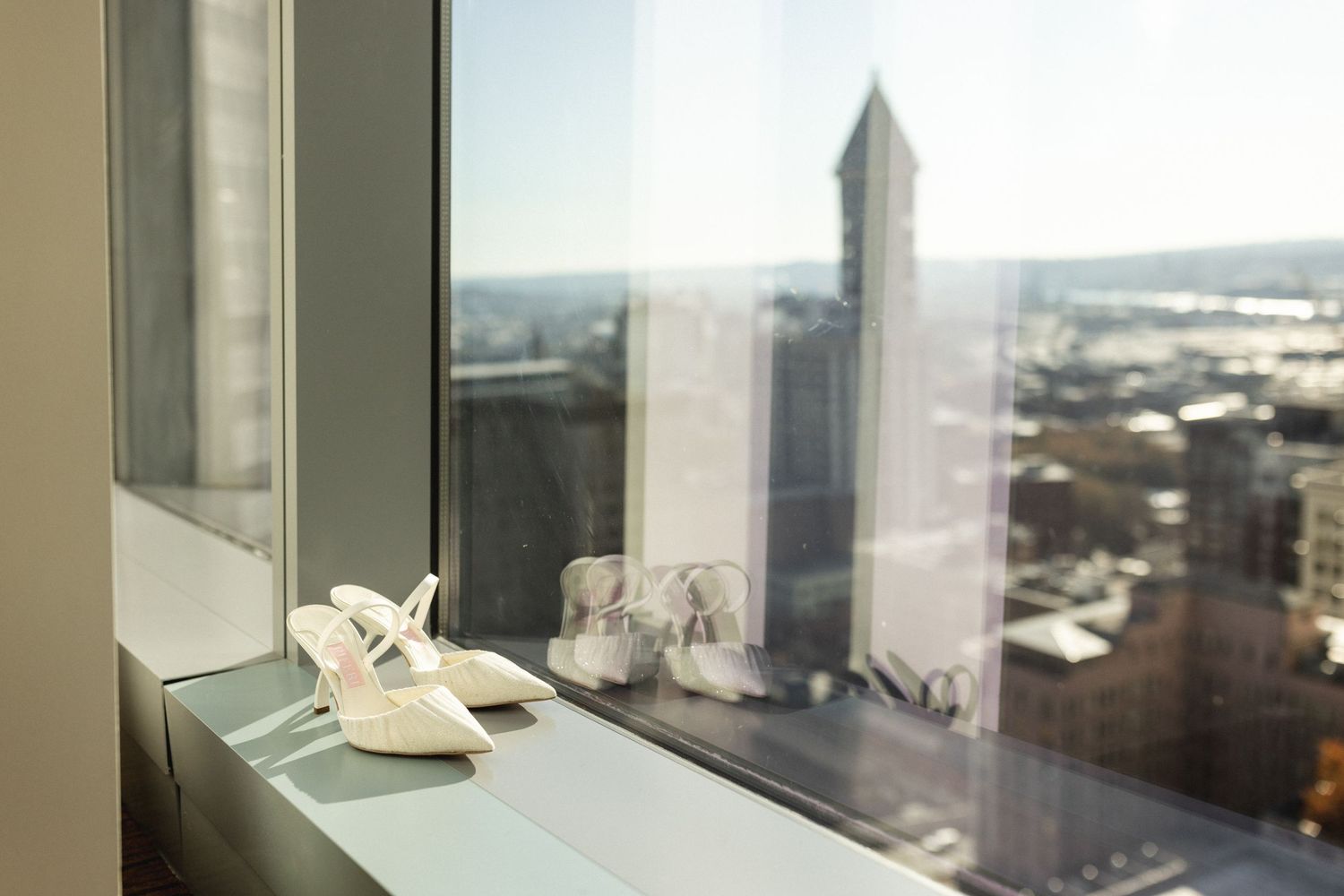 White bridal heels displayed on a high-rise window ledge with a city skyline view.