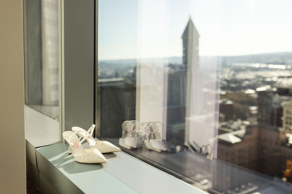 White bridal heels displayed on a high-rise window ledge with a city skyline view.