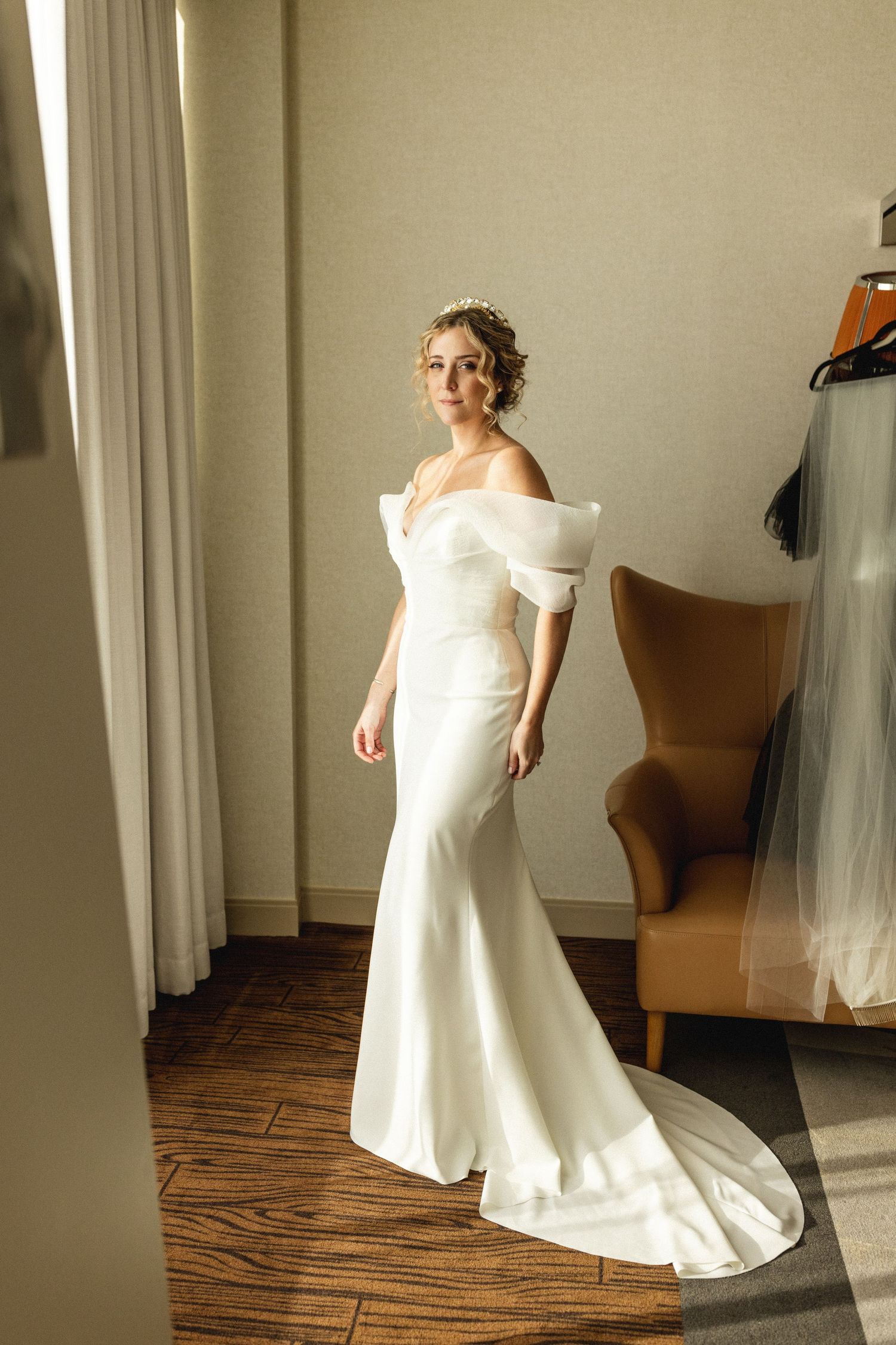 Elegant bride in an off-shoulder white mermaid wedding gown posing in a sunlit hotel room.