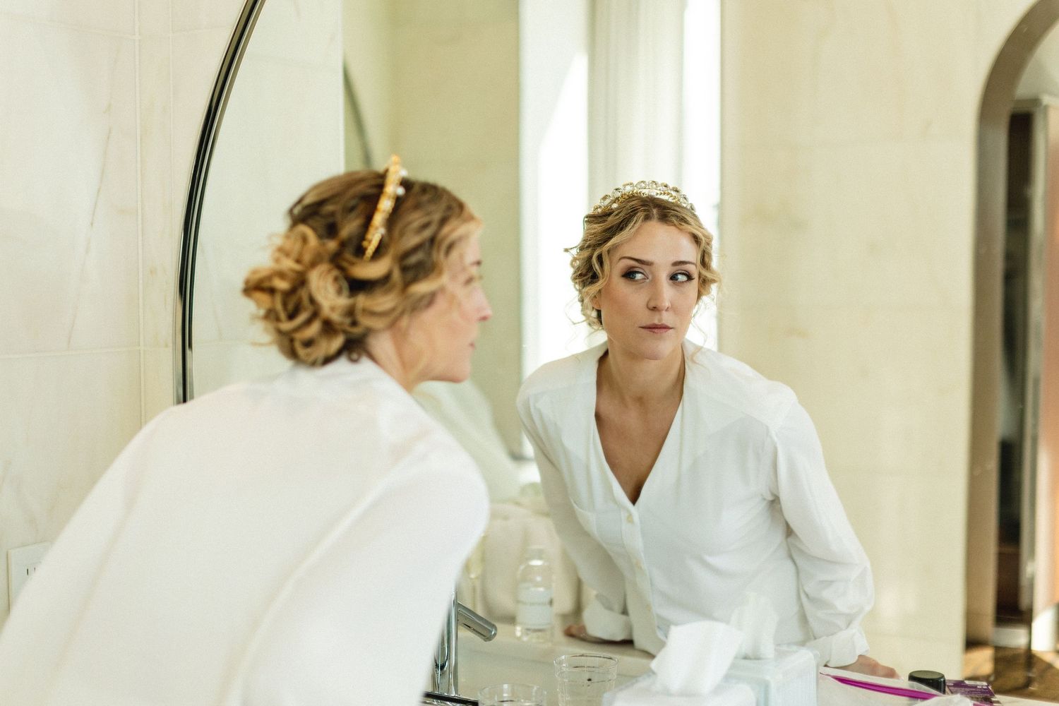 Bride in white robe examining her hair and makeup in a bathroom mirror before her wedding.