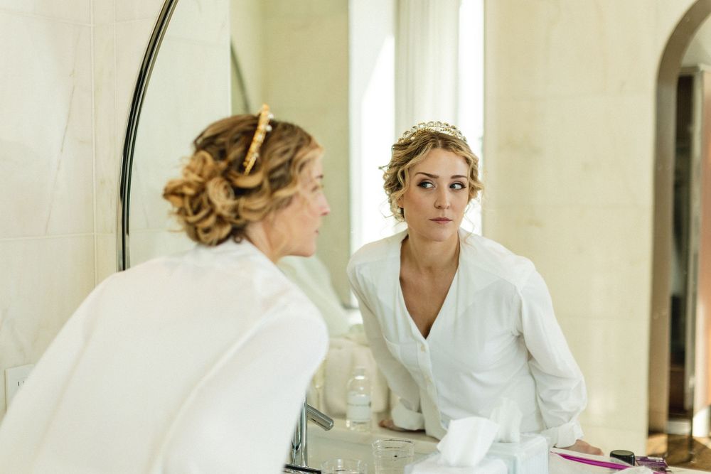 Bride in white robe examining her hair and makeup in a bathroom mirror before her wedding.