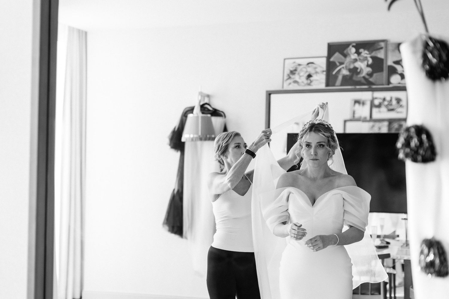 Black and white photo of a bridesmaid helping the bride put on her veil in a hotel room.
