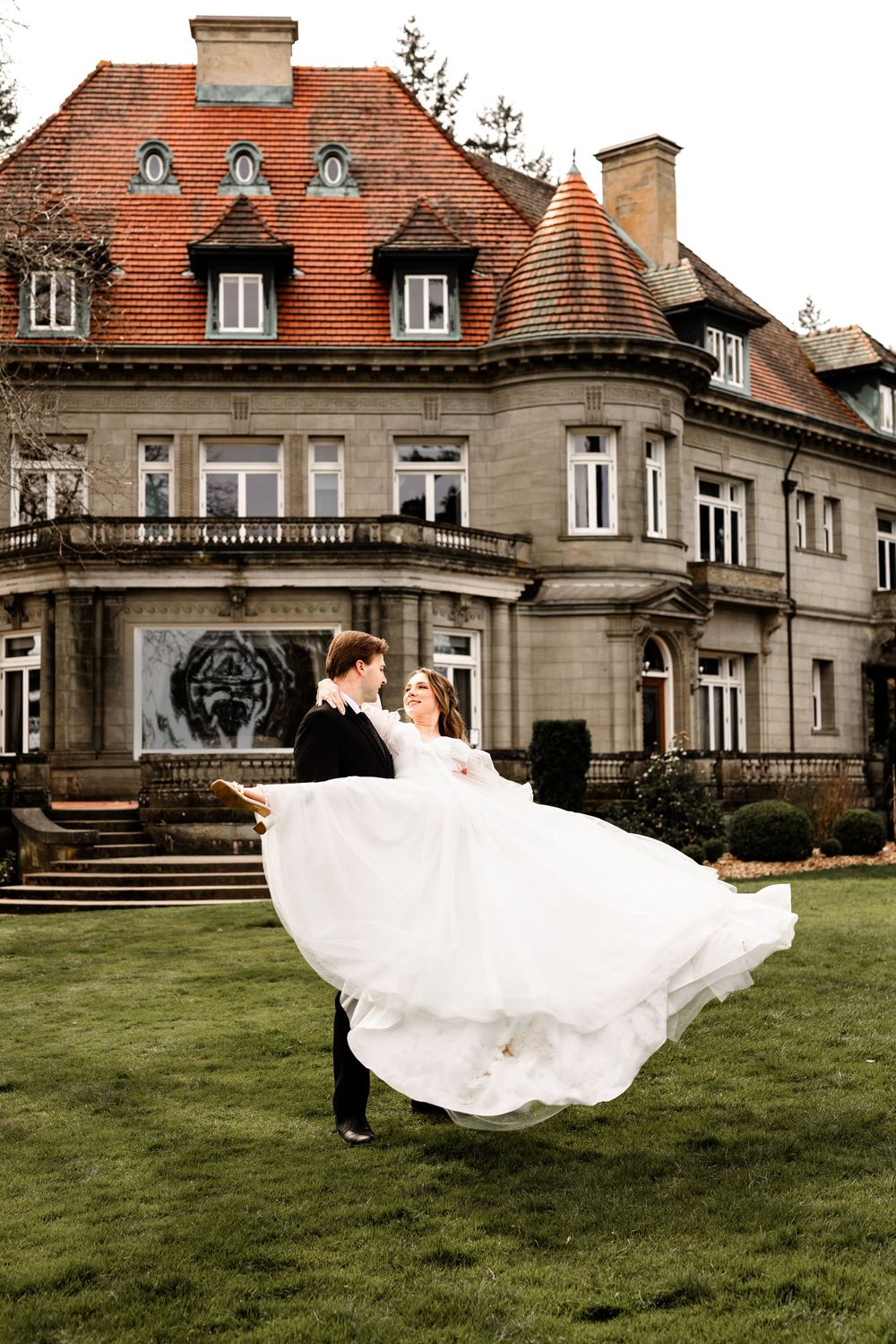 Groom lifting bride in white gown outside elegant stone mansion with red roof on green lawn.