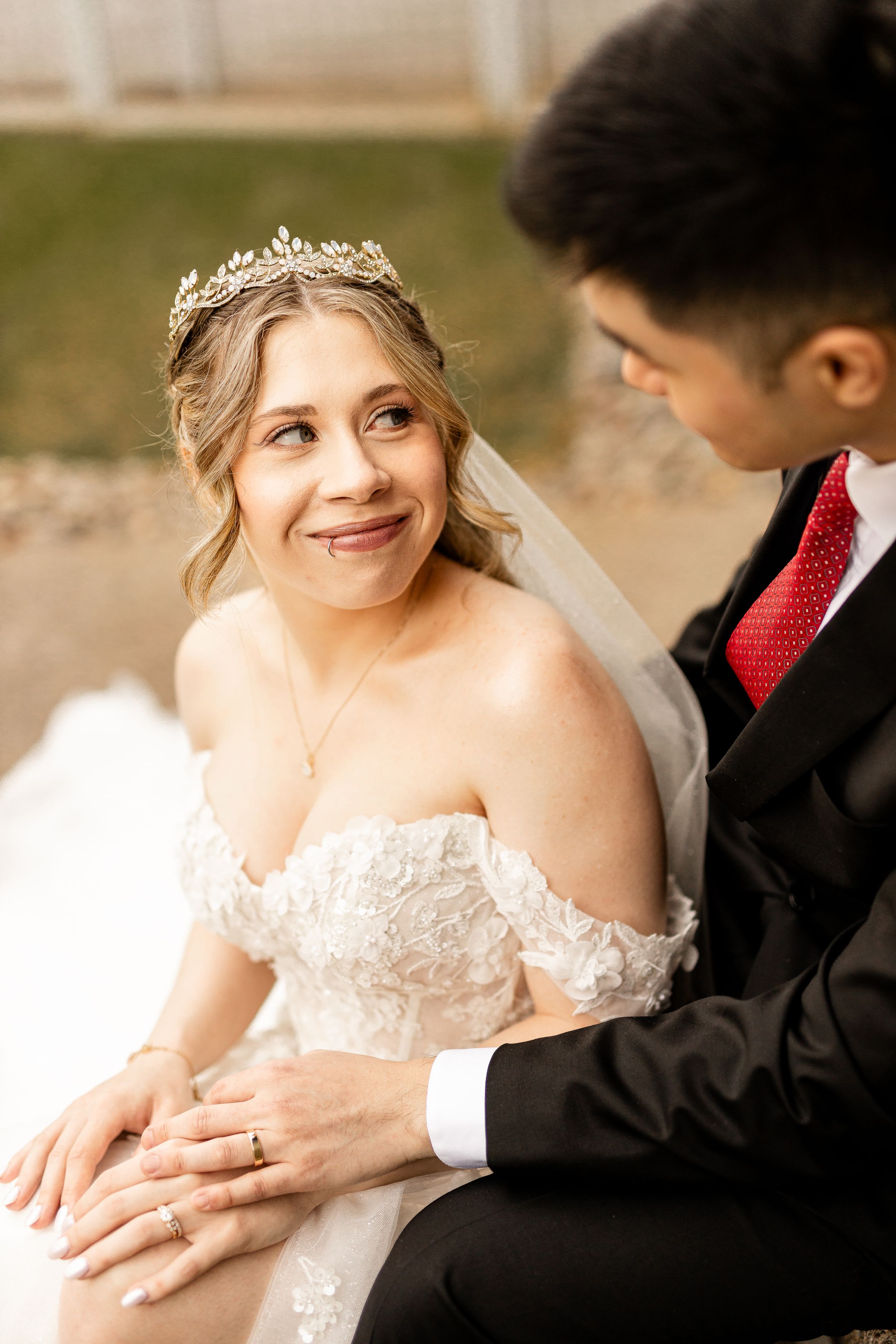 Bride in lace off-shoulder gown and gold tiara smiles lovingly at groom in black suit with red tie.