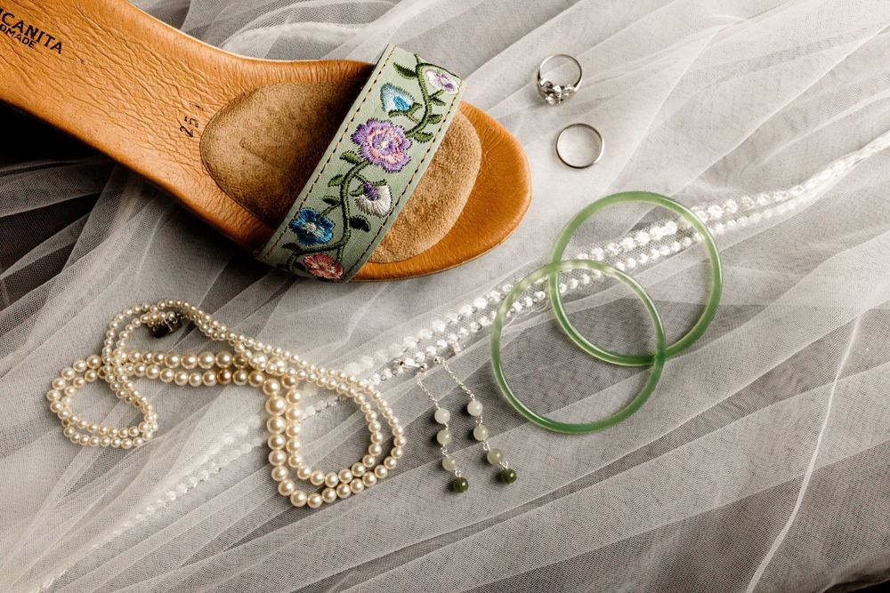 Bridal accessories flat lay featuring pearl necklace, green bangles, ring, earrings, and floral embroidered sandal on white tulle.