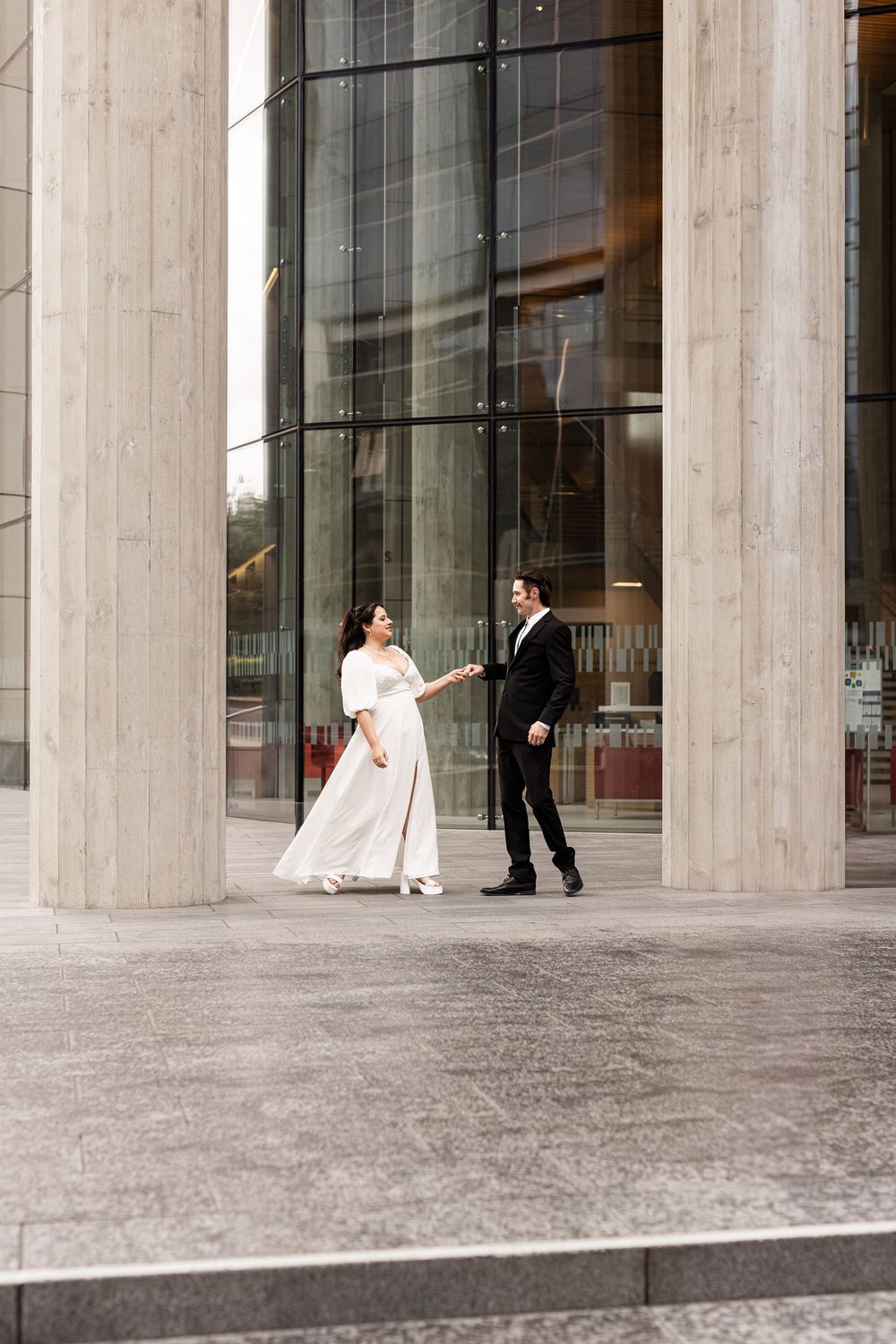 Bride in white gown and groom in black suit holding hands outside a modern building with tall columns.