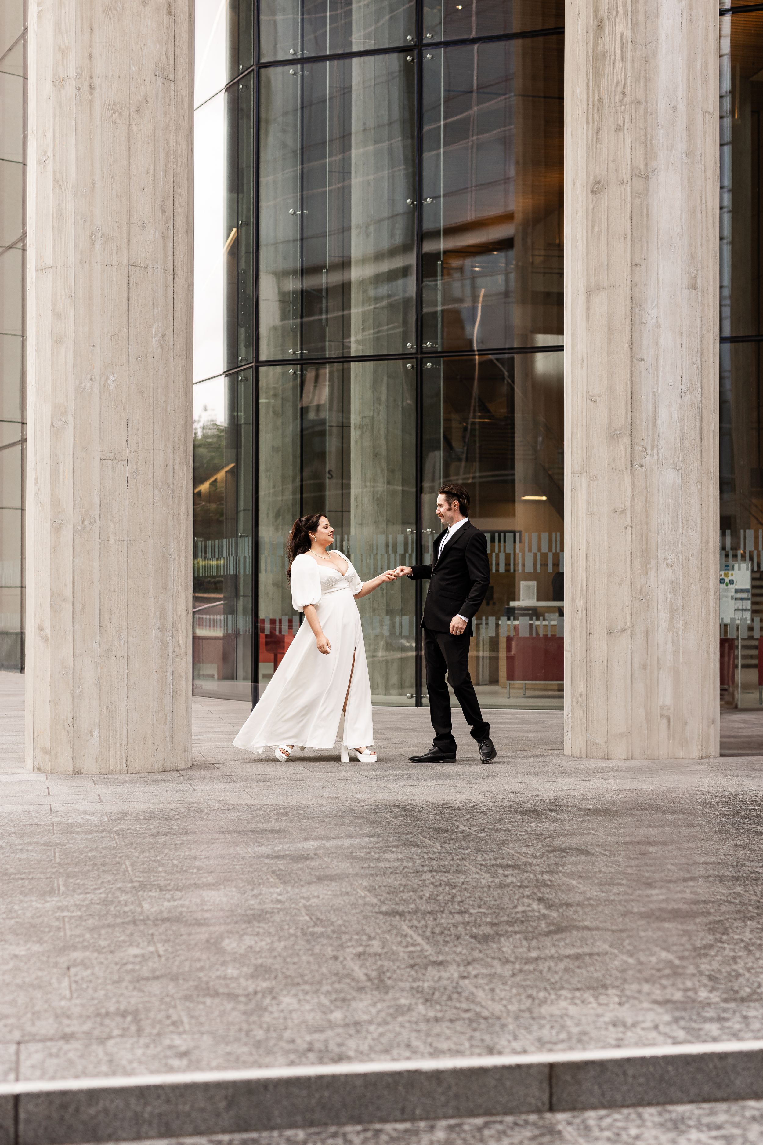 Bride in white gown and groom in black suit holding hands outside a modern building with tall columns.